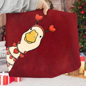 May include: A burgundy pillow featuring a whimsical illustration of a goose adorned with heart-shaped decorations. The goose has a yellow beak and is wearing a headband with heart accents. The design is set against a solid burgundy background.