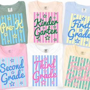 Puede incluir: Seis camisetas de colores con diseños de nivel escolar. Las camisetas son verdes, melocotón, azules, rosas, blancas y amarillas. Cada una tiene un diseño de rayas verticales con estrellas y el nivel escolar impreso en la parte delantera.