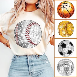 Puede incluir: Una camiseta blanca con un gráfico de béisbol brillante. La pelota de béisbol es blanca con costuras rojas y una textura brillante. Hay otras cuatro imágenes de diferentes pelotas de deporte con texturas brillantes: una pelota de baloncesto, una pelota de softbol, una pelota de fútbol y una pelota de voleibol.