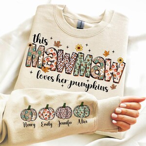 Puede incluir: Una sudadera beige con el texto "This Mawmaw loves her pumpkins" en letras de colores con patrones. La sudadera tiene cuatro peque&ntilde;as calabazas en la manga con los nombres "Henry", "Emily", "Jennifer" y "Alice" debajo.