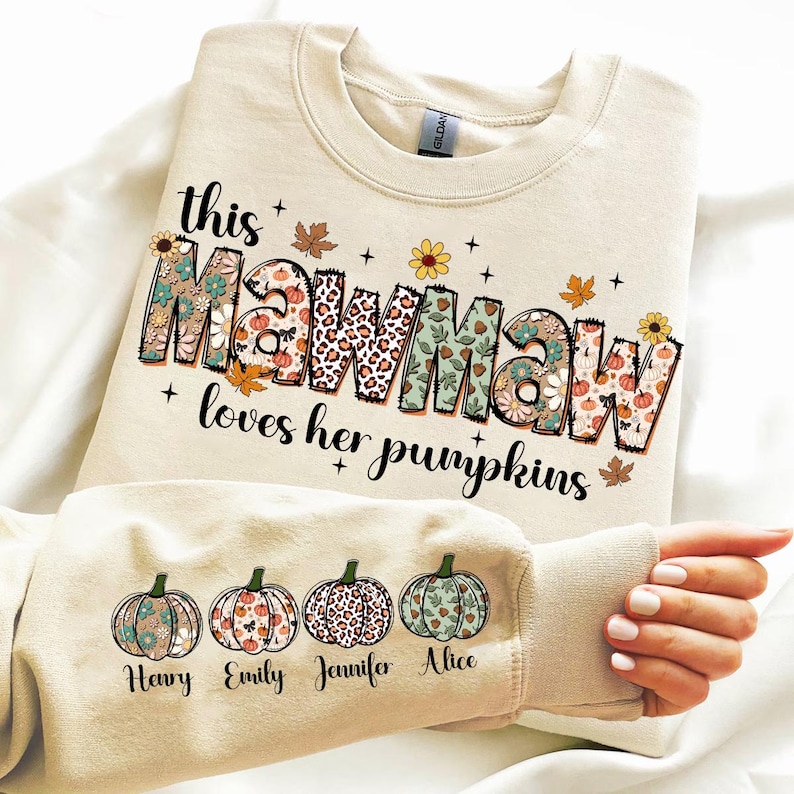 Puede incluir: Una sudadera beige con el texto "This Mawmaw loves her pumpkins" en letras de colores con patrones. La sudadera tiene cuatro peque&ntilde;as calabazas en la manga con los nombres "Henry", "Emily", "Jennifer" y "Alice" debajo.