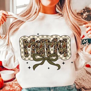 May include: White sweatshirt featuring the word "Mama" in a camouflage design, framed by a checkered pattern. A green bow sits beneath the word, with small black star accents.