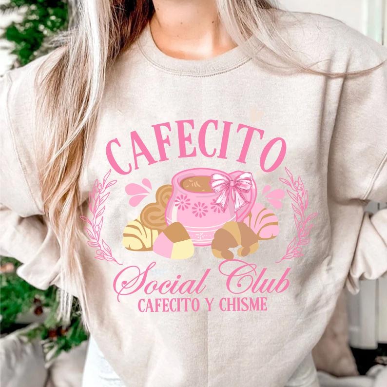 May include: A light beige sweatshirt with a pink graphic design that says "Cafecito Social Club Cafecito y Chisme". The graphic features a pink coffee mug with a pink bow, surrounded by pink and brown pastries.