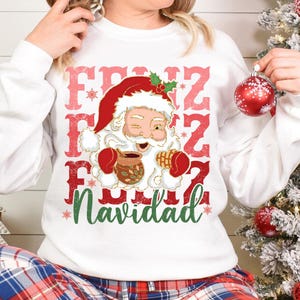 May include: White sweatshirt with a red and green Christmas design featuring a cartoon Santa Claus holding a mug and the text "Feliz Navidad".
