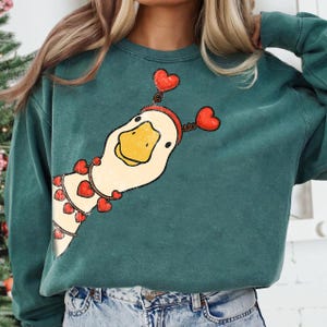May include: Teal green sweatshirt featuring a whimsical goose design. The goose is adorned with red heart-shaped accents, including a headband and a garland. The design is printed on a comfortable, casual-fit sweatshirt.
