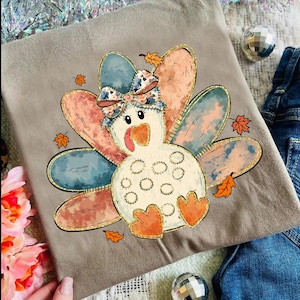 May include: A tan-colored t-shirt featuring a cartoon turkey design. The turkey has a white body, orange feet and beak, and a patterned bow. The turkey's feathers are in shades of blue, pink, and gold, with autumn leaves scattered around.