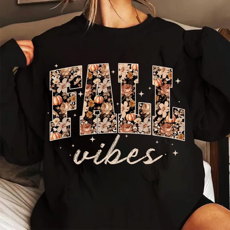 May include: Black sweatshirt with the word "FALL" in large, floral-filled letters, featuring pumpkins and flowers. Below, the word "vibes" is written in a cursive font. White star accents surround the text.