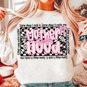 Peut inclure: Sweat-shirt blanc avec le mot "Motherhood" en rose, orn&eacute; de n&oelig;uds roses et de marguerites. Le motif est plac&eacute; sur un fond &agrave; carreaux noir et blanc. Les phrases "Some days I rock it. Some days it rocks me" sont &eacute;galement imprim&eacute;es sur le v&ecirc;tement.