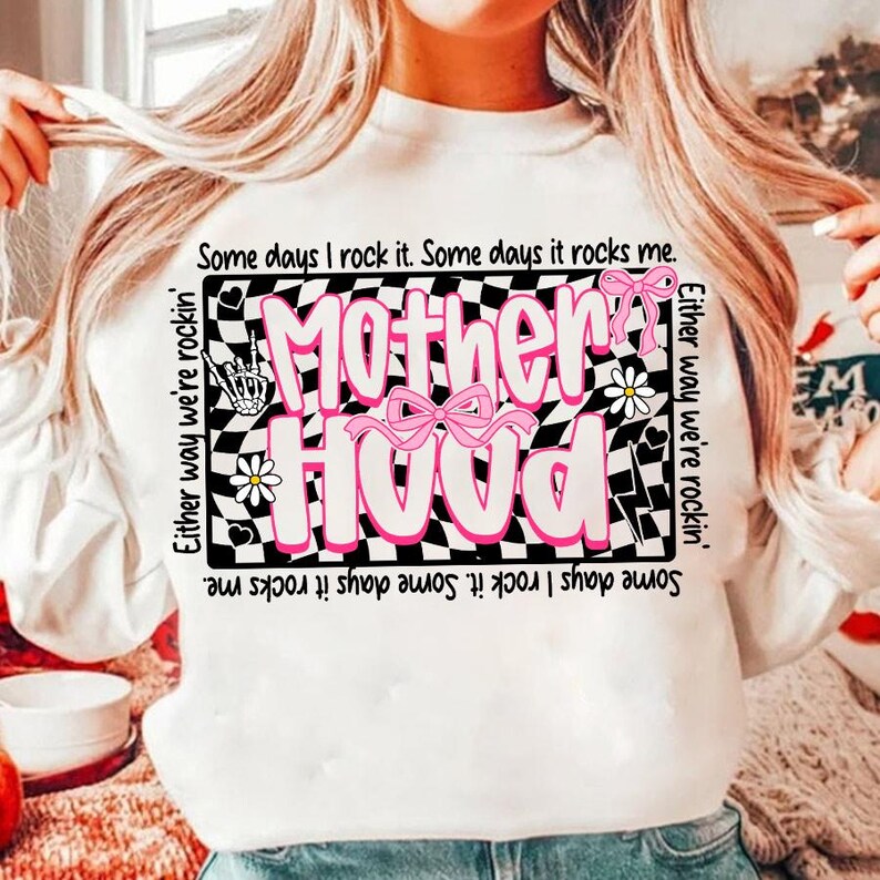 Peut inclure: Sweat-shirt blanc avec le mot "Motherhood" en rose, orn&eacute; de n&oelig;uds roses et de marguerites. Le motif est plac&eacute; sur un fond &agrave; carreaux noir et blanc. Les phrases "Some days I rock it. Some days it rocks me" sont &eacute;galement imprim&eacute;es sur le v&ecirc;tement.