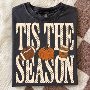 May include: A charcoal gray short-sleeve t-shirt featuring the text TIS THE SEASON in large, cream-colored, polka-dotted letters. Between the two lines of text are a brown football, an orange pumpkin, and a tan coffee cup with a lid.
