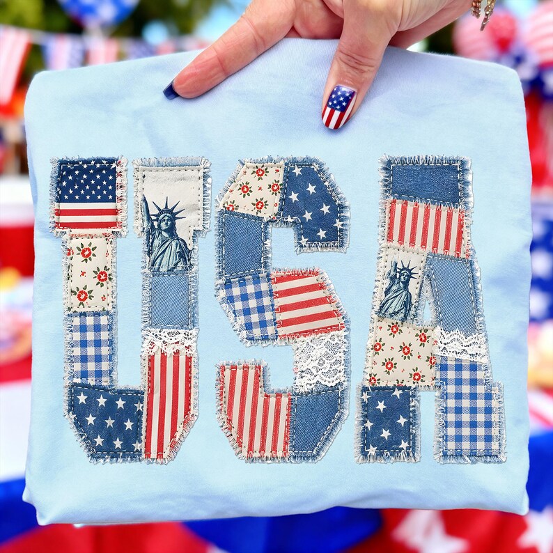 K&ouml;nnte beinhalten: Hellblaues T-Shirt mit dem Wort "USA" in einem Patchwork-Design appliziert. Die Buchstaben zeigen verschiedene patriotische Muster, darunter die amerikanische Flagge, Sterne, Streifen und Blumendrucke. Das T-Shirt wird von einer Hand mit passendem Nageldesign gehalten.