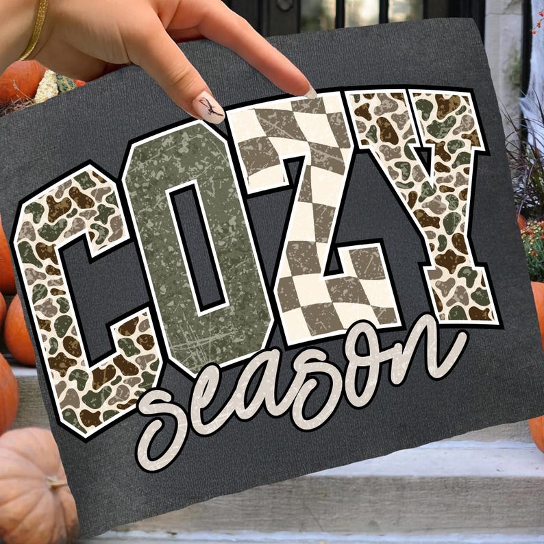 K&ouml;nnte beinhalten: Dunkelgraues T-Shirt mit dem Schriftzug "COZY Season" in einem Used-Look-Design. Die Buchstaben haben ein Tarn-, Schachbrett- und olivgr&uuml;nes Muster. Das Shirt wird mit K&uuml;rbissen im Hintergrund pr&auml;sentiert.