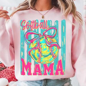 May include: Pink sweatshirt with a graphic featuring a softball with a bow, and the words "Softball Mama" in bright colors. The design includes teal and yellow accents, perfect for a softball mom.
