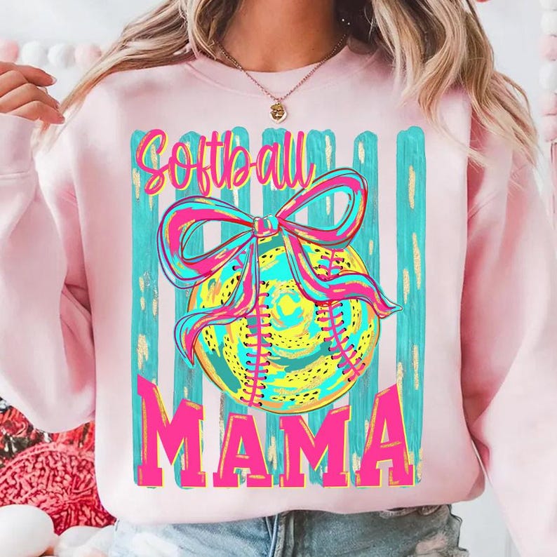 May include: Pink sweatshirt with a graphic featuring a softball with a bow, and the words "Softball Mama" in bright colors. The design includes teal and yellow accents, perfect for a softball mom.