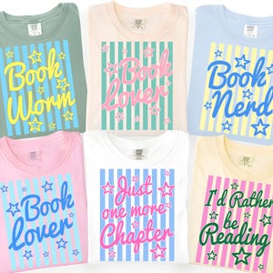 May include: Six t-shirts in various colors, each with a unique book-themed graphic. The shirts feature phrases like "Book Worm," "Book Lover," and "I'd Rather be Reading," with star and stripe designs. The shirts are folded and displayed.
