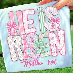 May include: Light blue t-shirt with the words "HE IS RISEN" in large, colorful letters. The letters are decorated with pink, green, and patterned fabrics, and accented with bows. The text "Matthew 28:6" is written below in pink.
