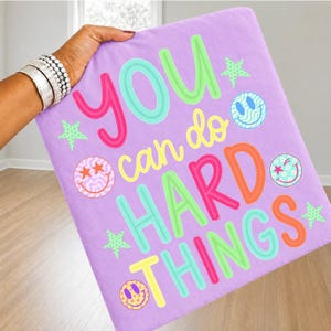 May include: A square lavender pillowcase featuring the colorful text YOU can do HARD THINGS in shades of pink, yellow, green, and orange. The design includes several small green stars and three circular smiley face icons with various patterns.