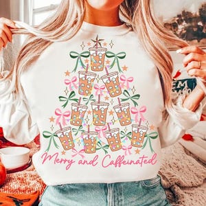 Puede incluir: Sudadera blanca con un diseño de árbol de Navidad hecho de vasos de café helado. El árbol está decorado con lazos rosas, guirnaldas verdes y luces de colores. El texto "Merry and Caffeinated" está impreso debajo del árbol.