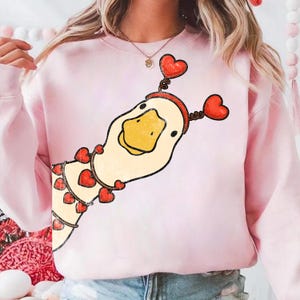 May include: A light pink sweatshirt featuring a cartoon duck with a yellow beak and a red heart-shaped headband. The duck is decorated with red hearts. The sweatshirt is a Valentine's Day or love-themed design.