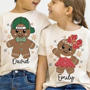 May include: Two white t-shirts featuring gingerbread people designs. The shirt on the left has a green hat and says "David". The shirt on the right has a red bow and says "Emily".