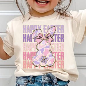May include: A white t-shirt with a pink and purple bunny rabbit graphic. The bunny has a pink bow with the words "Egg Hunter" on it. The text "Happy Easter" is repeated in pink and purple letters.
