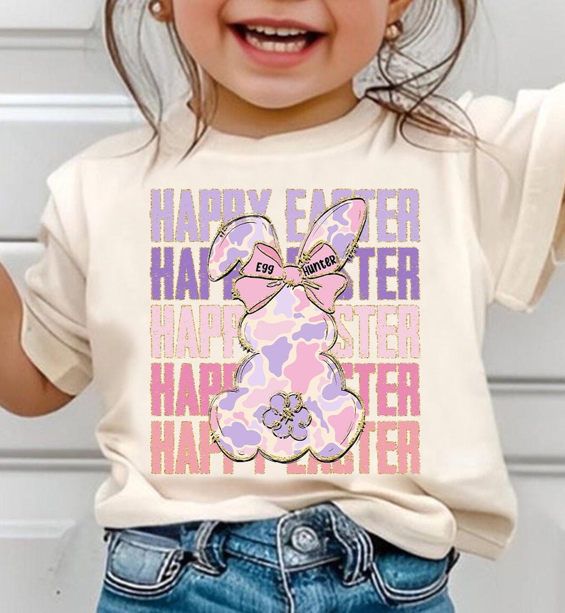 May include: A white t-shirt with a pink and purple bunny rabbit graphic. The bunny has a pink bow with the words "Egg Hunter" on it. The text "Happy Easter" is repeated in pink and purple letters.