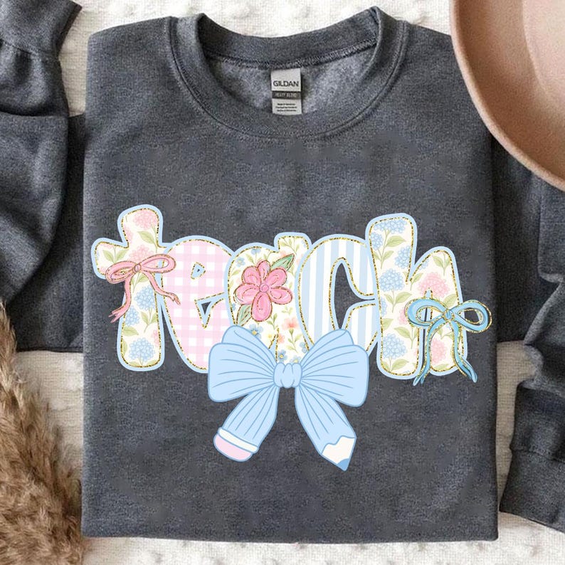 Puede incluir: Una sudadera gris con un dise&ntilde;o de patchwork que deletrea "Teach" con un lazo azul y un l&aacute;piz.