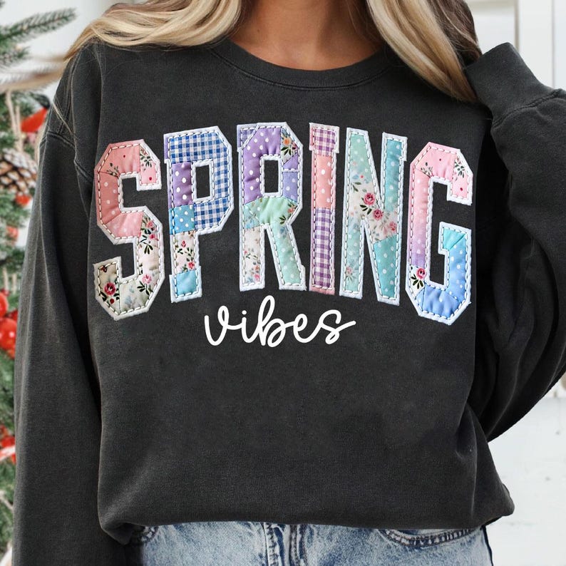 May include: Dark gray sweatshirt with the words "SPRING VIBES" in a patchwork design. The letters are in various pastel colors and floral patterns, with a white outline. The word "vibes" is in a cursive font.