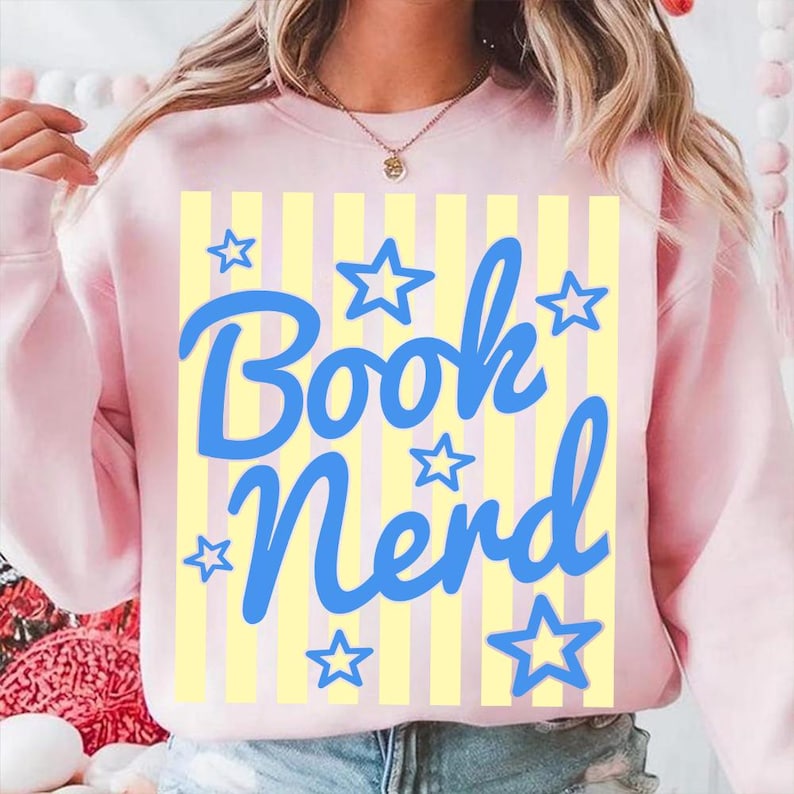 May include: A light pink sweatshirt with the words "Book Nerd" in blue script, surrounded by blue star shapes. The background features vertical yellow stripes. A casual, comfortable top for book lovers.