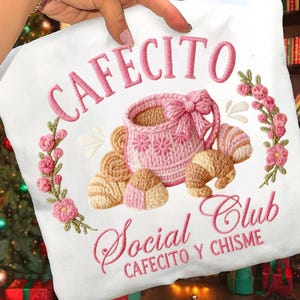 Pode incluir: Saco tote branco com bordado rosa. O design inclui a palavra "CAFECITO" acima de uma ch&aacute;vena de caf&eacute; rosa com um la&ccedil;o, rodeada por flores e pastelaria. As palavras "Social Club" e "CAFECITO Y CHISME" est&atilde;o em baixo.