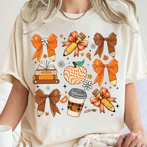 May include: A white t-shirt with a colorful design featuring bows, pencils, books, a coffee cup, and an apple. The design is in shades of orange, brown, and white.