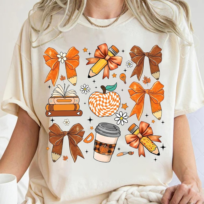 May include: A white t-shirt with a colorful design featuring bows, pencils, books, a coffee cup, and an apple. The design is in shades of orange, brown, and white.