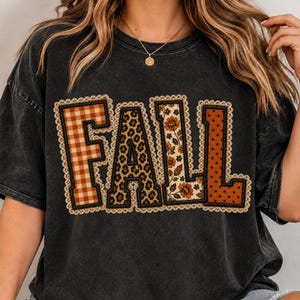 May include: A charcoal gray short-sleeve t-shirt featuring the word FALL in large block letters. Each letter has a unique pattern: orange gingham, leopard print, floral print, and orange polka dots, all outlined with a cream lace trim.