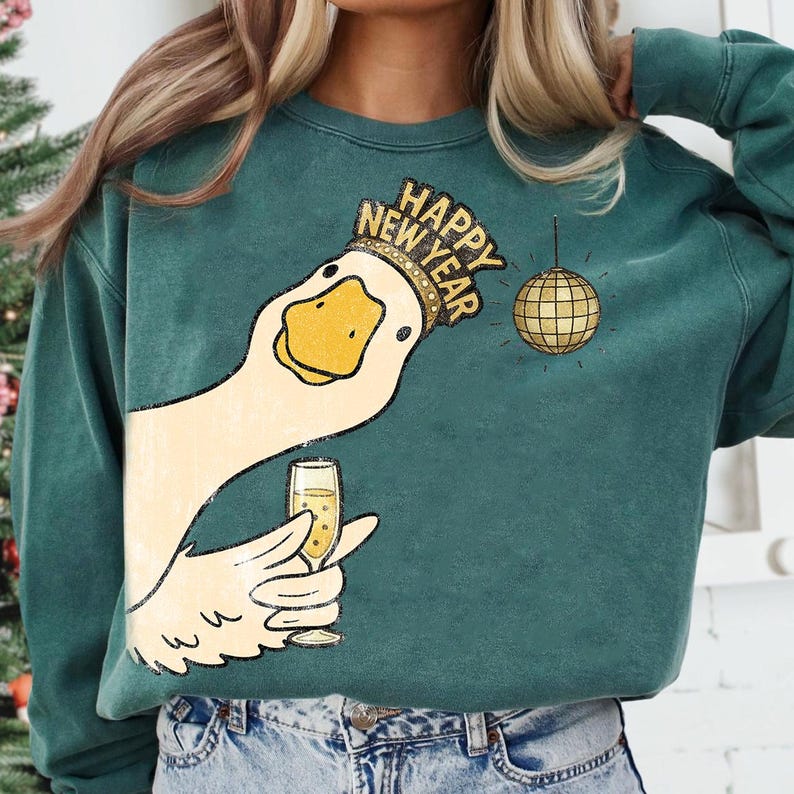 May include: Teal green sweatshirt with a graphic of a goose wearing a crown that says "Happy New Year." The goose is holding a champagne glass, and a gold disco ball hangs nearby. The sweatshirt is a relaxed fit.