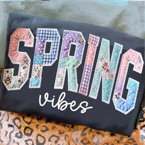 May include: Dark gray t-shirt with the word "SPRING" in patchwork letters, featuring floral and polka dot patterns. The word "vibes" is written in white script below the word "SPRING".
