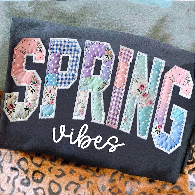 May include: Dark gray t-shirt with the word "SPRING" in patchwork letters, featuring floral and polka dot patterns. The word "vibes" is written in white script below the word "SPRING".