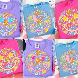 May include: Six colorful t-shirts in shades of purple, blue, and pink, each featuring a large smiley face design. The smiley faces are decorated with stars, hearts, and lightning bolts, surrounded by floral and abstract patterns. The shirts are folded and displayed.