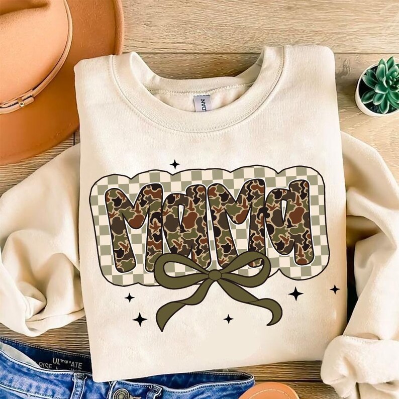 May include: A cream-coloured sweatshirt featuring the word "MAMA" in a camouflage pattern, outlined with a chequered design. A green bow sits below the word, with black star accents. The sweatshirt is on a wooden surface.