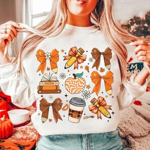 May include: White sweatshirt with a colorful autumn-themed design featuring bows, pencils, books, a coffee cup, and an apple. The design is in shades of orange, brown, and yellow.