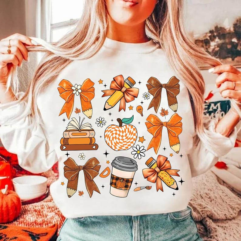 May include: White sweatshirt with a colorful autumn-themed design featuring bows, pencils, books, a coffee cup, and an apple. The design is in shades of orange, brown, and yellow.