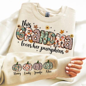 Puede incluir: Una sudadera beige con el texto "This Grandma loves her pumpkins" en letras de colores con patrones. Cuatro peque&ntilde;as calabazas con dise&ntilde;os florales est&aacute;n en la manga con los nombres "Henry", "Emily", "Jennifer" y "Alice" debajo.