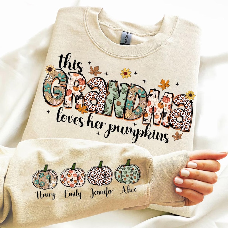 Puede incluir: Una sudadera beige con el texto "This Grandma loves her pumpkins" en letras de colores con patrones. Cuatro peque&ntilde;as calabazas con dise&ntilde;os florales est&aacute;n en la manga con los nombres "Henry", "Emily", "Jennifer" y "Alice" debajo.