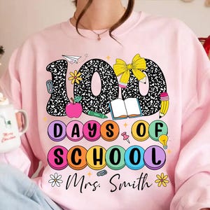 Puede incluir: Sudadera rosa con un diseño gráfico de los números 100 en blanco y negro con una textura de cuaderno. Los números están rodeados de círculos coloridos que dicen "Days of School" y un lazo, un libro, un lápiz y una bombilla. El texto "Mrs. Smith" está en la parte inferior del diseño.