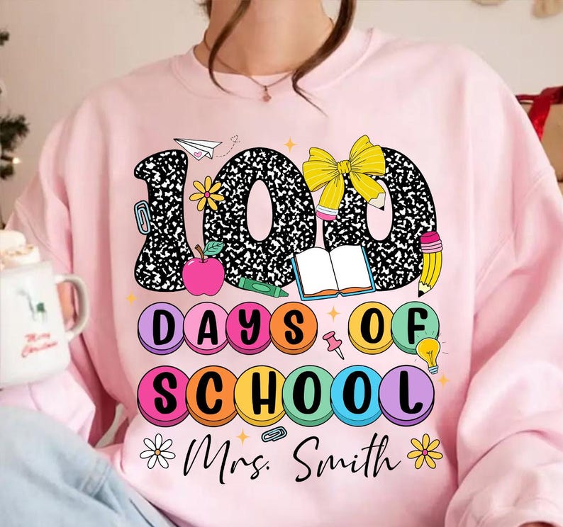 May include: Pink sweatshirt featuring a graphic design of the numbers 100 in black and white with a notebook texture. The numbers are surrounded by colourful circles that say "Days of School" and a bow, book, pencil, and lightbulb. The text "Mrs. Smith" is at the bottom of the design.