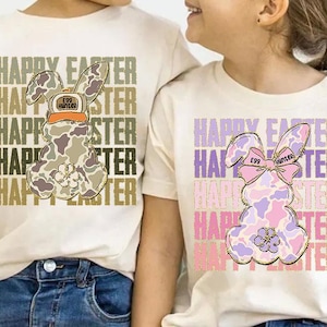 May include: Two white t-shirts with a camouflage bunny rabbit design. The bunny on the left is wearing a hat that says "Egg Hunter". The bunny on the right is wearing a pink bow that says "Egg Hunter". Both shirts have the text "Happy Easter" repeated multiple times.