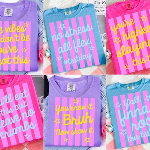 May include: A collection of six colorful t-shirts with various phrases and star graphics. The shirts are in shades of purple, blue, and pink. Each shirt has a unique message in a vertical striped design, such as "the vibes don't lie you've got this."