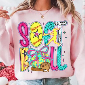 May include: Pink sweatshirt with a colorful graphic design that says "Softball" with a softball, bat, glove, and a player hitting a ball.