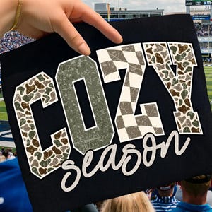 K&ouml;nnte beinhalten: Schwarzes T-Shirt mit den Worten "COZY season" in wei&szlig;er Schrift. Die Buchstaben sind wei&szlig; umrandet und mit einem Tarnmuster und Schachbrettmuster versehen. Das T-Shirt wird von einer Hand vor einem Stadion hochgehalten.