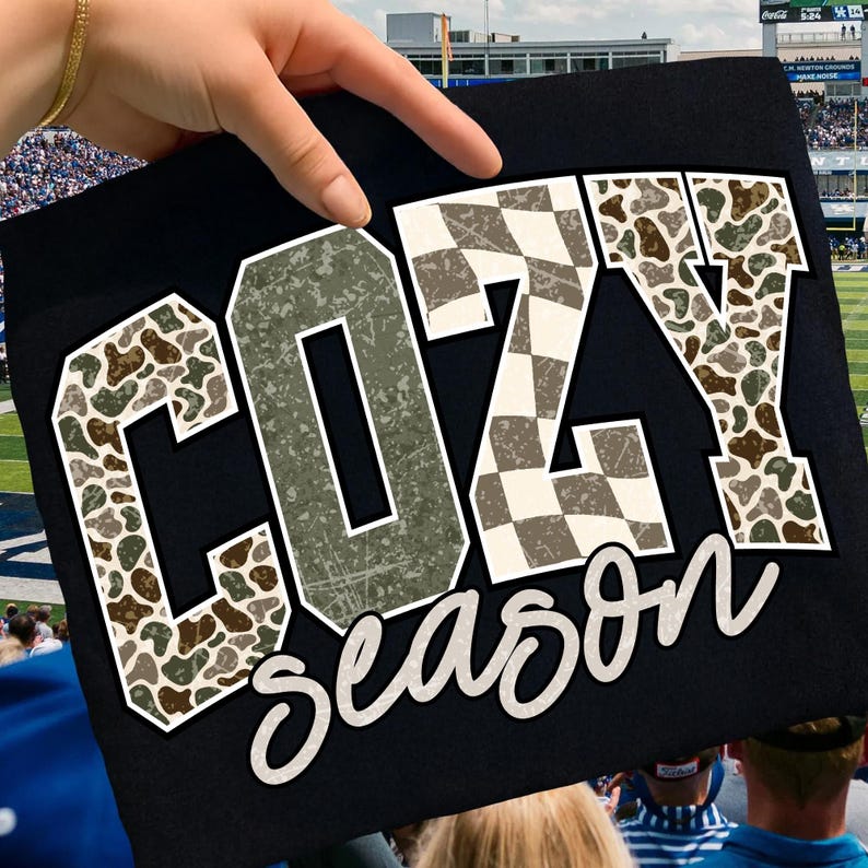 K&ouml;nnte beinhalten: Schwarzes T-Shirt mit den Worten "COZY season" in wei&szlig;er Schrift. Die Buchstaben sind wei&szlig; umrandet und mit einem Tarnmuster und Schachbrettmuster versehen. Das T-Shirt wird von einer Hand vor einem Stadion hochgehalten.