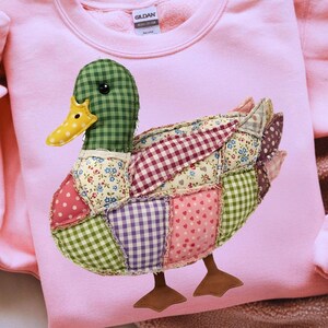 May include: A light pink sweatshirt features a patchwork duck design. The duck has a green checkered head, a yellow beak, and a body composed of various fabric patterns, including polka dots and floral prints. The duck's feet are brown.